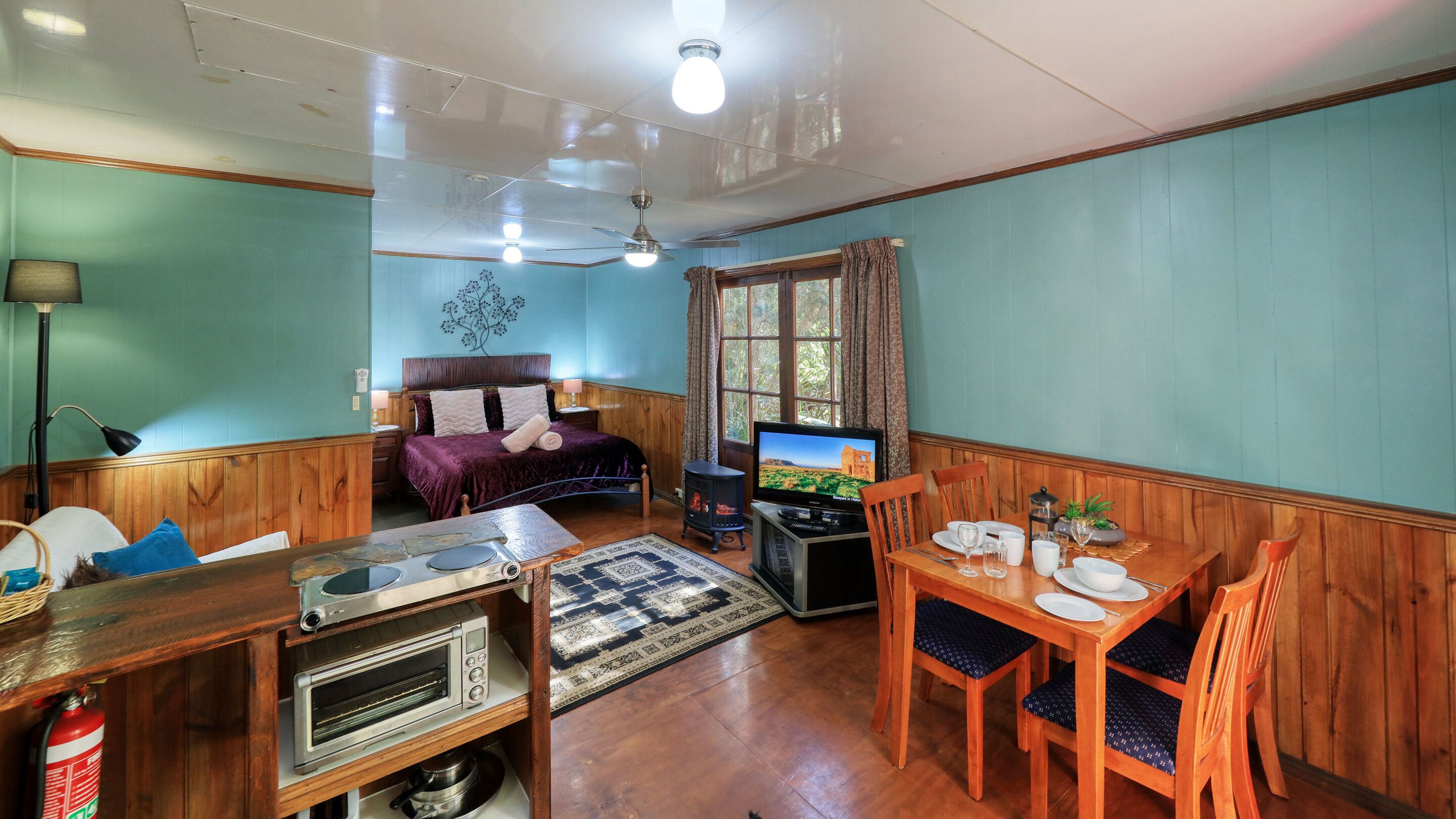 A cozy room with a bed, a television, a dining table with chairs, a small kitchen area with a stove and oven, and a window with curtains.