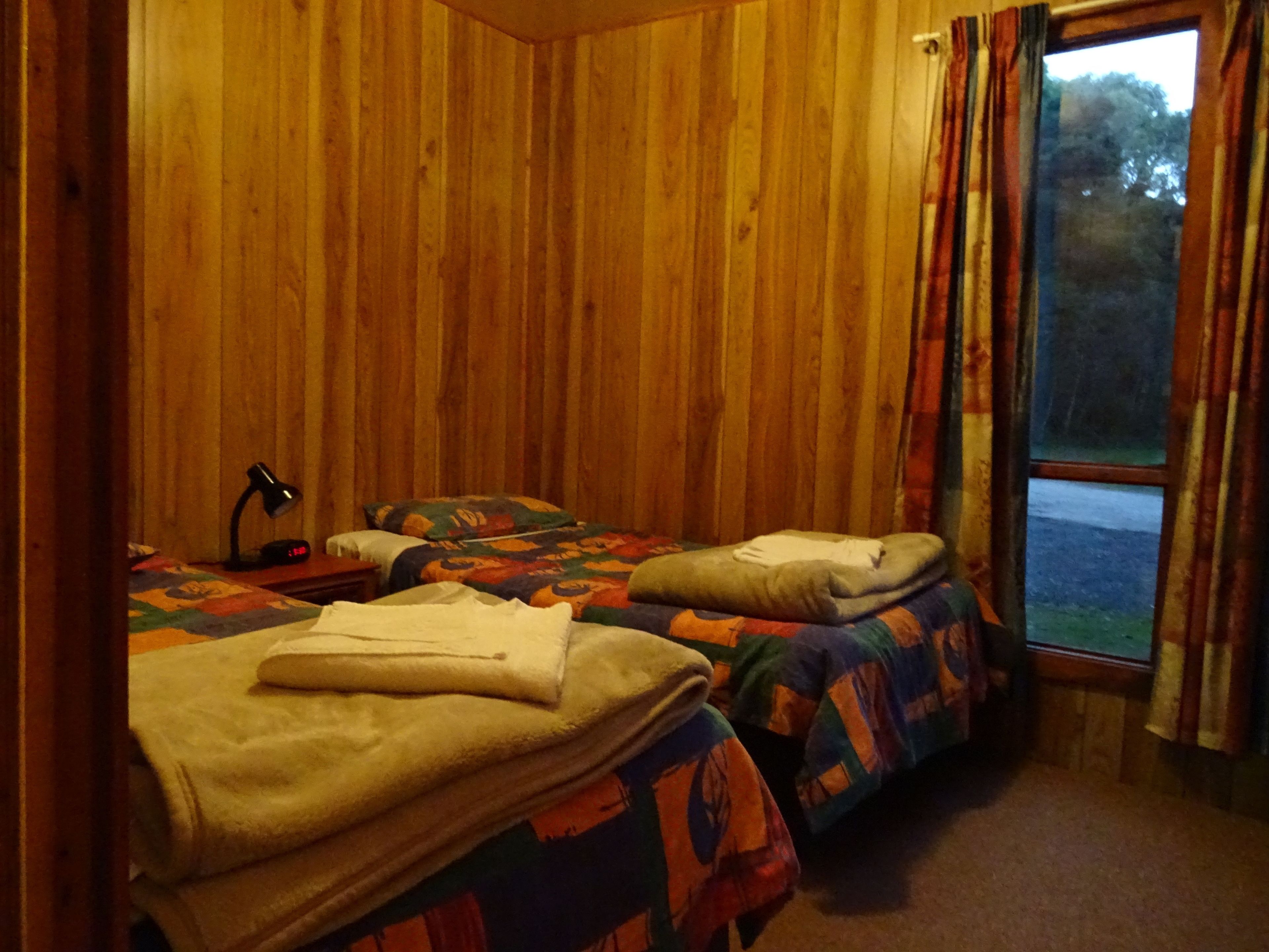A room with two beds, wooden walls, a desk with a lamp, and a window with curtains.