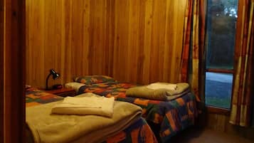 A room with two beds, wooden walls, a desk with a lamp, and a window with curtains.