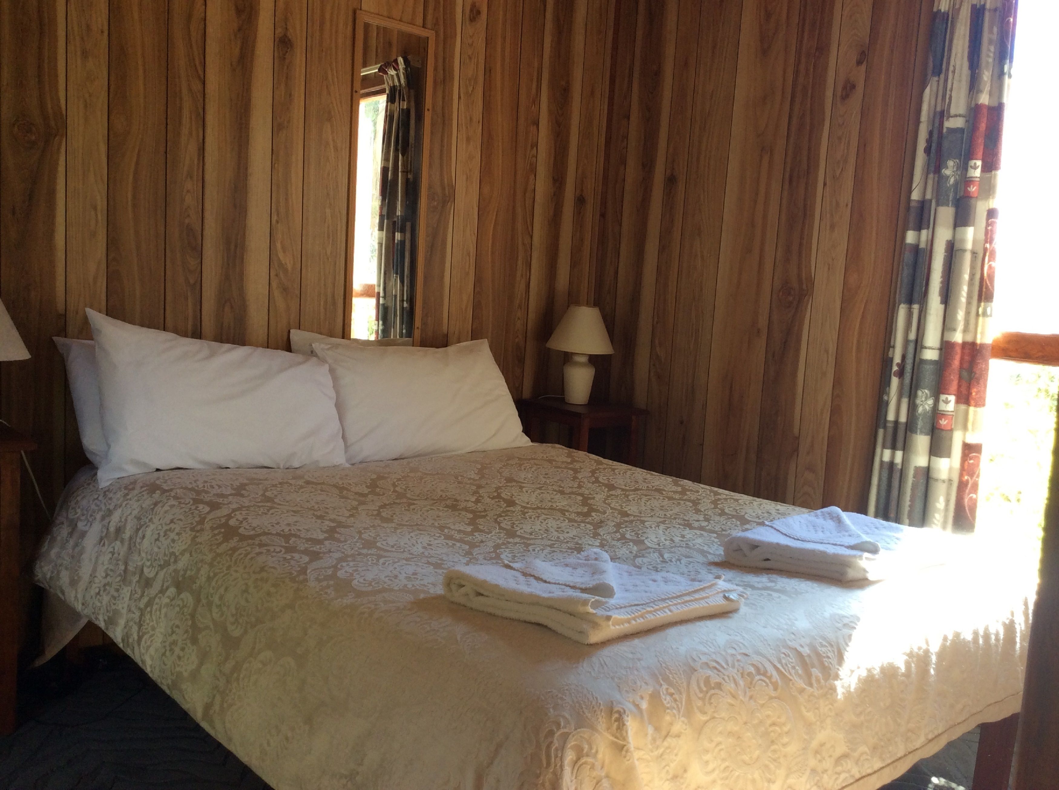Una habitación con paneles de madera, una cama, almohadas, una lámpara y un ventanal con cortinas.
