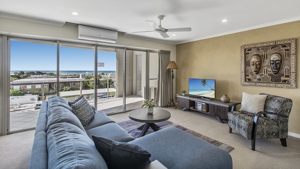 A modern living room with a large sofa, a coffee table, a TV, and a patterned armchair. - Rovera Apartments (Maroochydore)