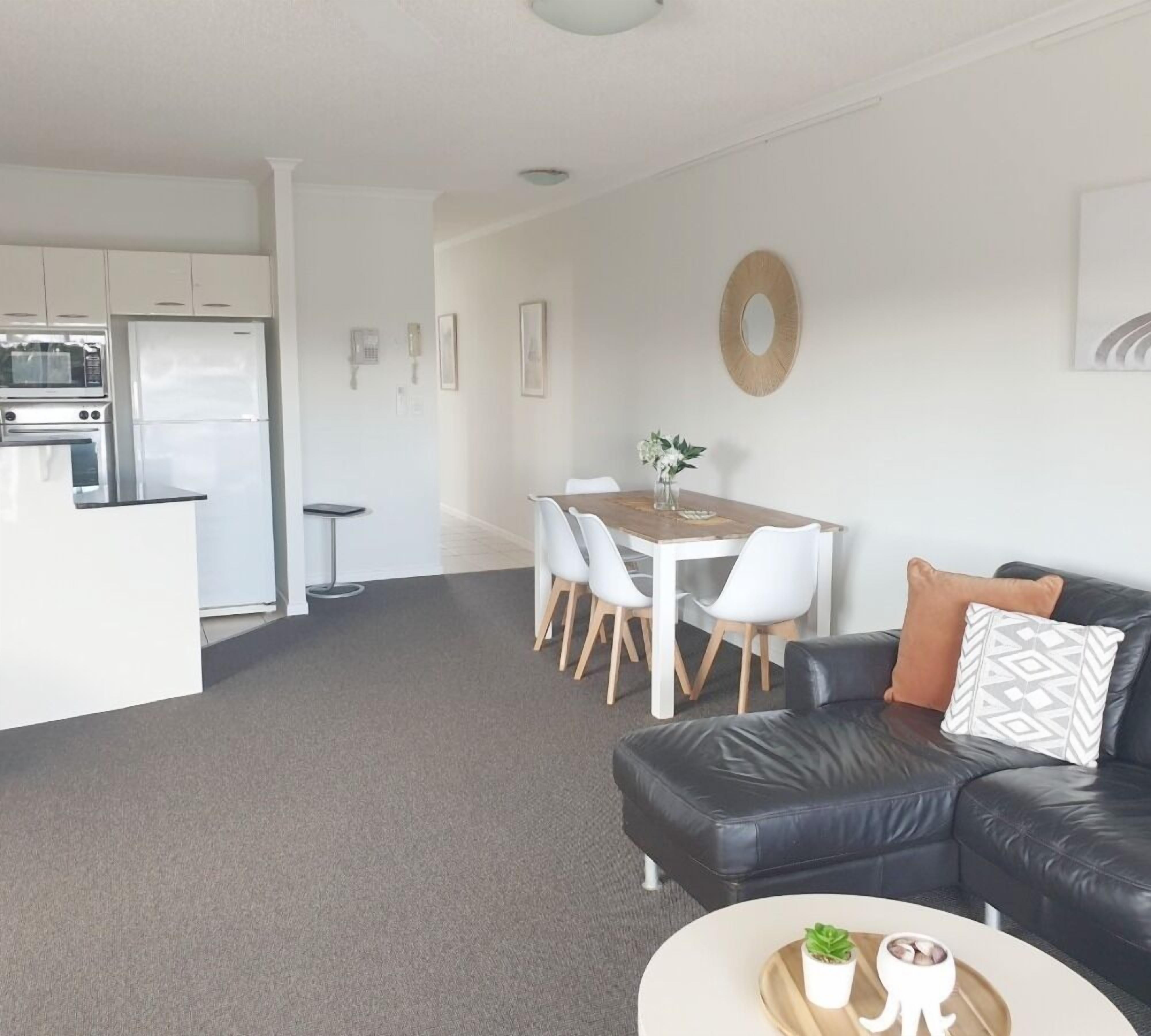 A modern living room with a black leather sofa, a dining table with white chairs, and a kitchen area with white appliances.