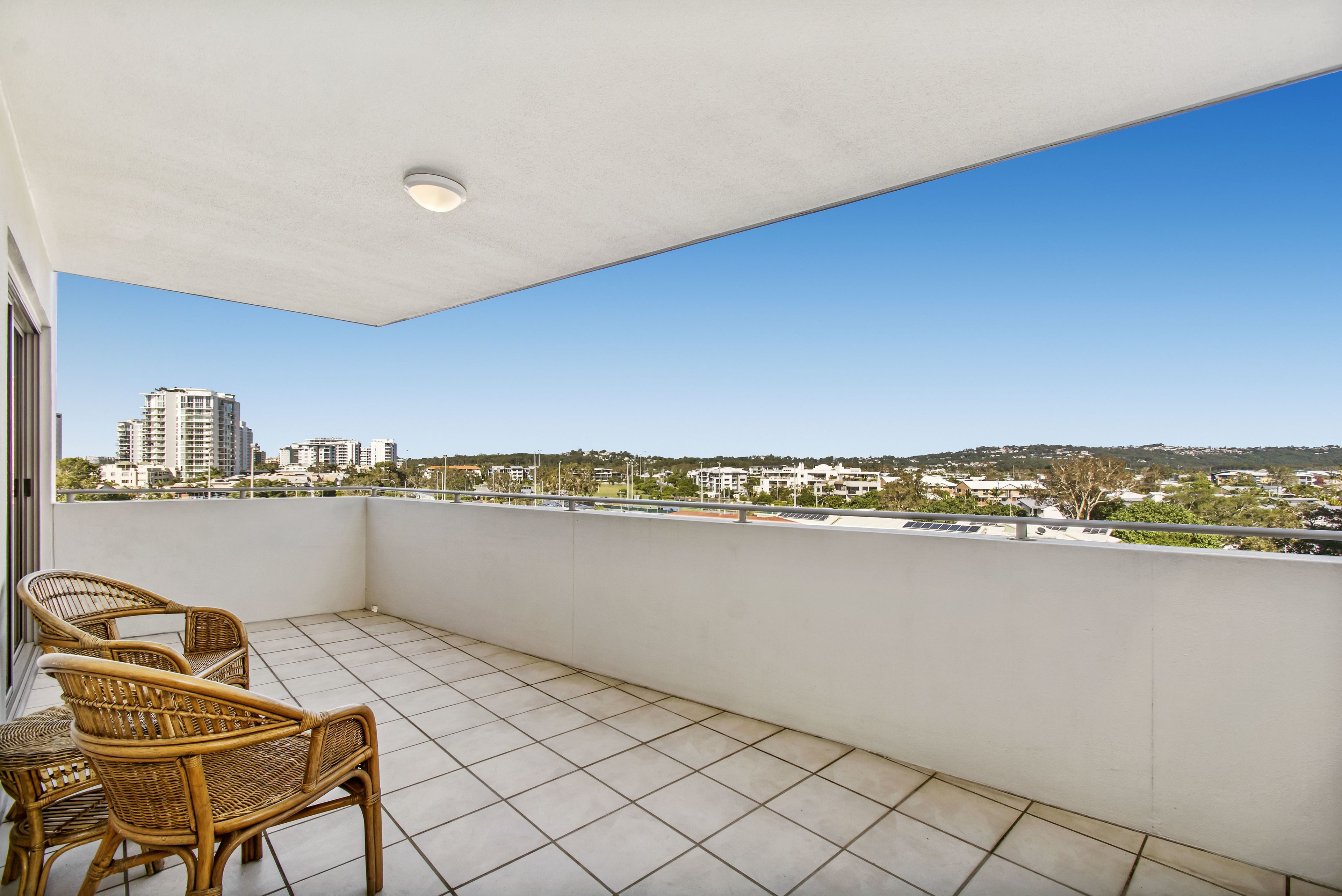 Un balcon doté d’un sol carrelé et de fauteuils en osier, offrant une vue sur un paysage urbain avec ses bâtiments et sa verdure.
