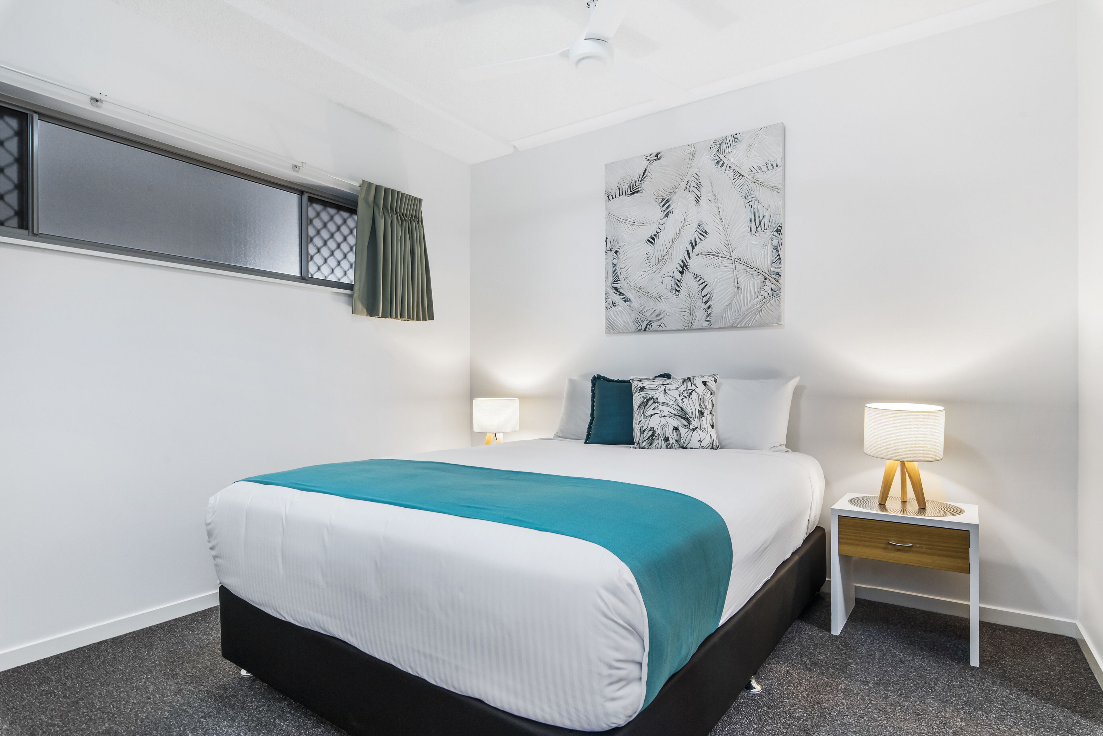 Una habitación de hotel con cama, dos mesitas de noche, un ventilador de techo, una ventana con cortina y una obra de arte colgada en la pared.