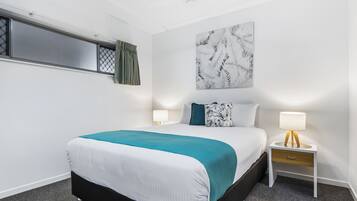 Una habitación de hotel con cama, dos mesitas de noche, un ventilador de techo, una ventana con cortina y una obra de arte colgada en la pared.