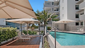 A poolside area with lounge chairs and umbrellas, a wooden deck, and a clear blue pool. - Rovera Apartments (Maroochydore)