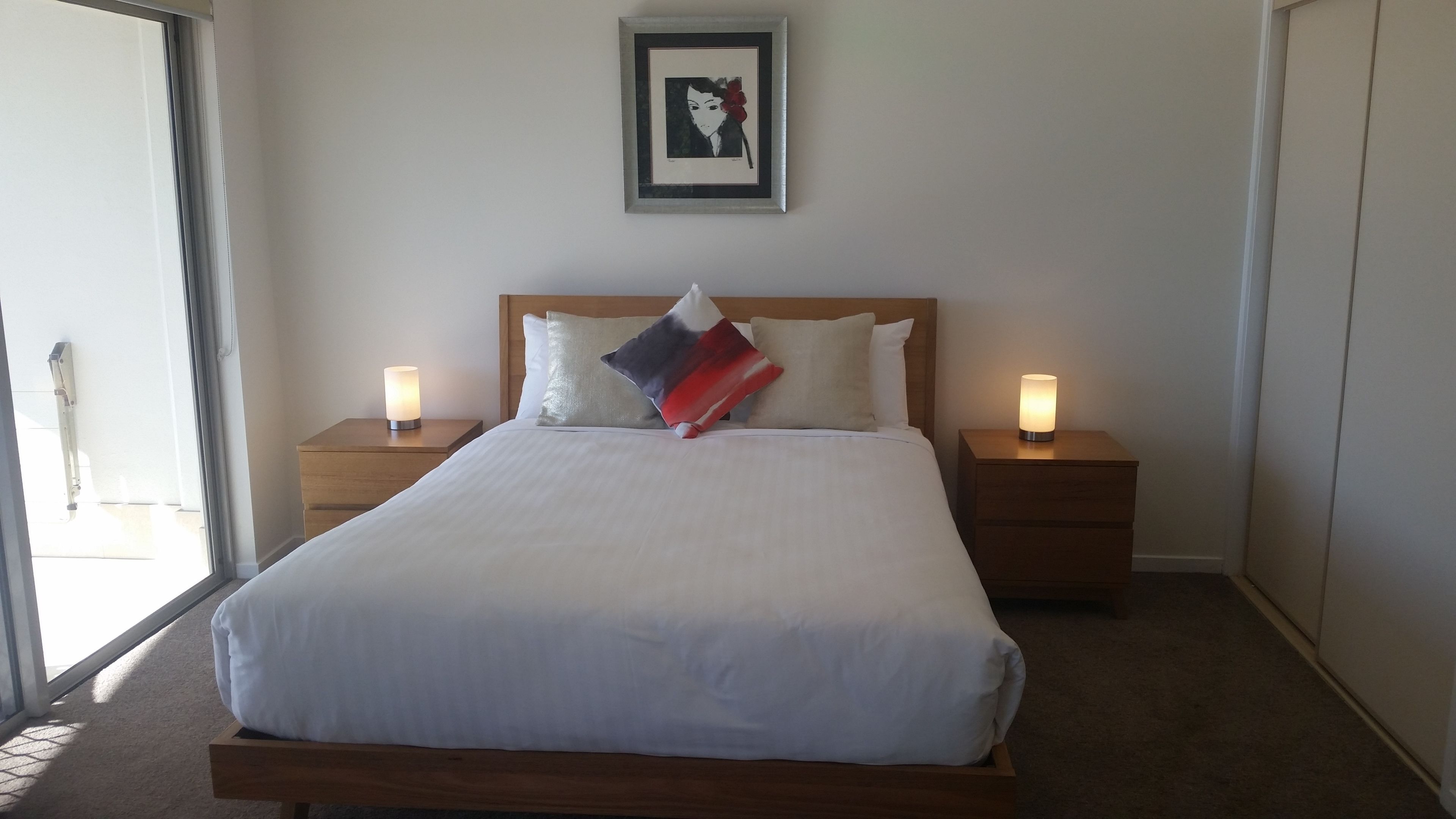 A neatly made bed with white linens, two bedside tables with lamps, a wooden headboard, and a framed artwork on the wall.