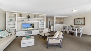 A modern living room with a flat-screen TV, a sofa, two armchairs, a coffee table, and a dining area with a table and chairs. - Rovera Apartments (Maroochydore)
