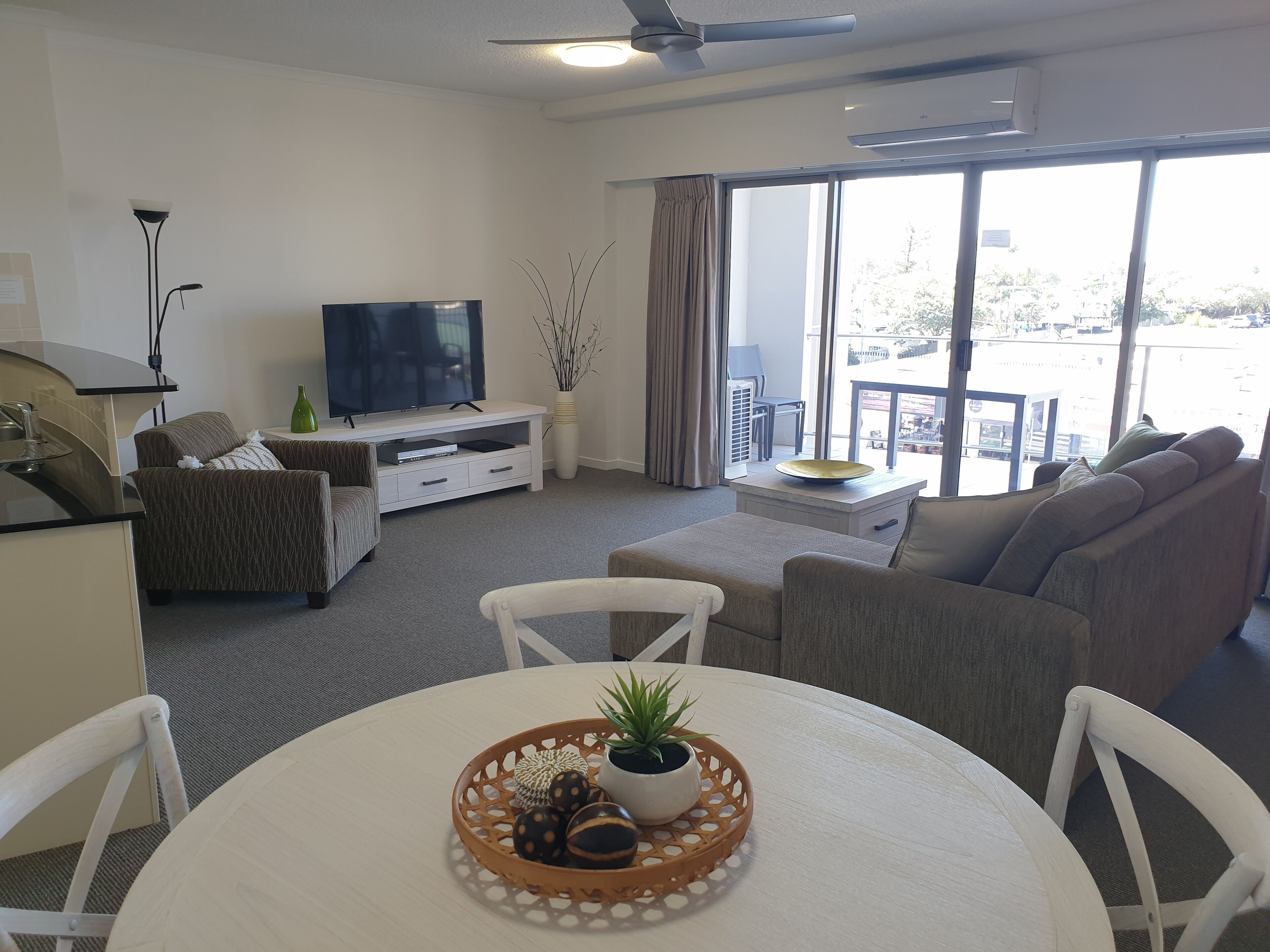 A modern living room with a flat-screen TV, sofa, dining table, and a balcony with a view.
