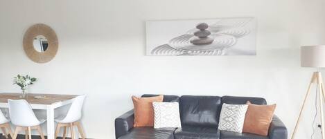A modern living room with a sectional sofa, a dining table with chairs, a ceiling fan, a floor lamp, and a wall-mounted mirror.