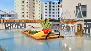 A glass table with a wooden board holding cheese, tomatoes, and herbs, and two glasses of wine on a balcony with buildings in the background. - Cityville Apartments & Motel (Rockhampton)