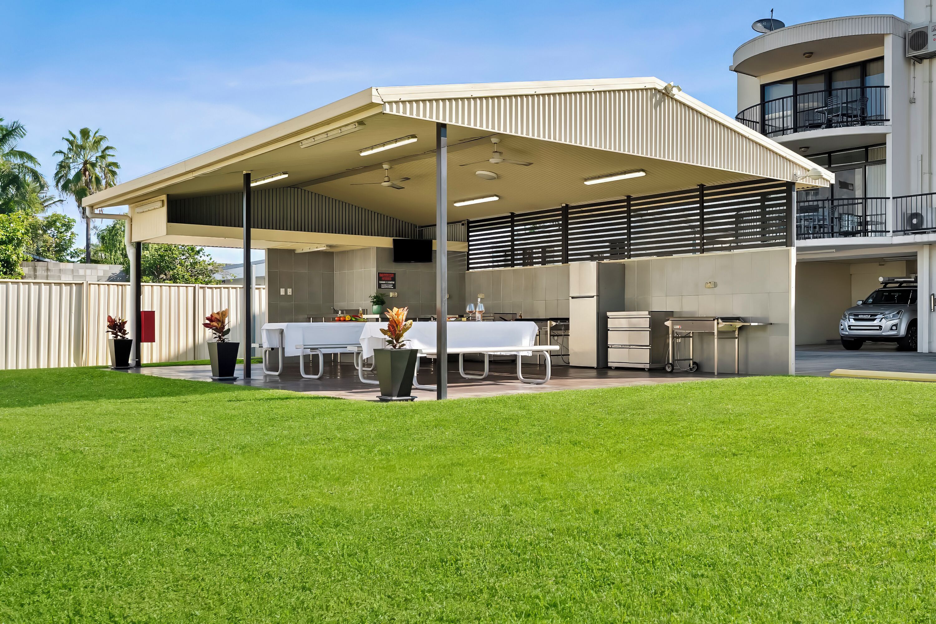 A covered outdoor dining area with tables and chairs, a grill, and a car parked in the background.
