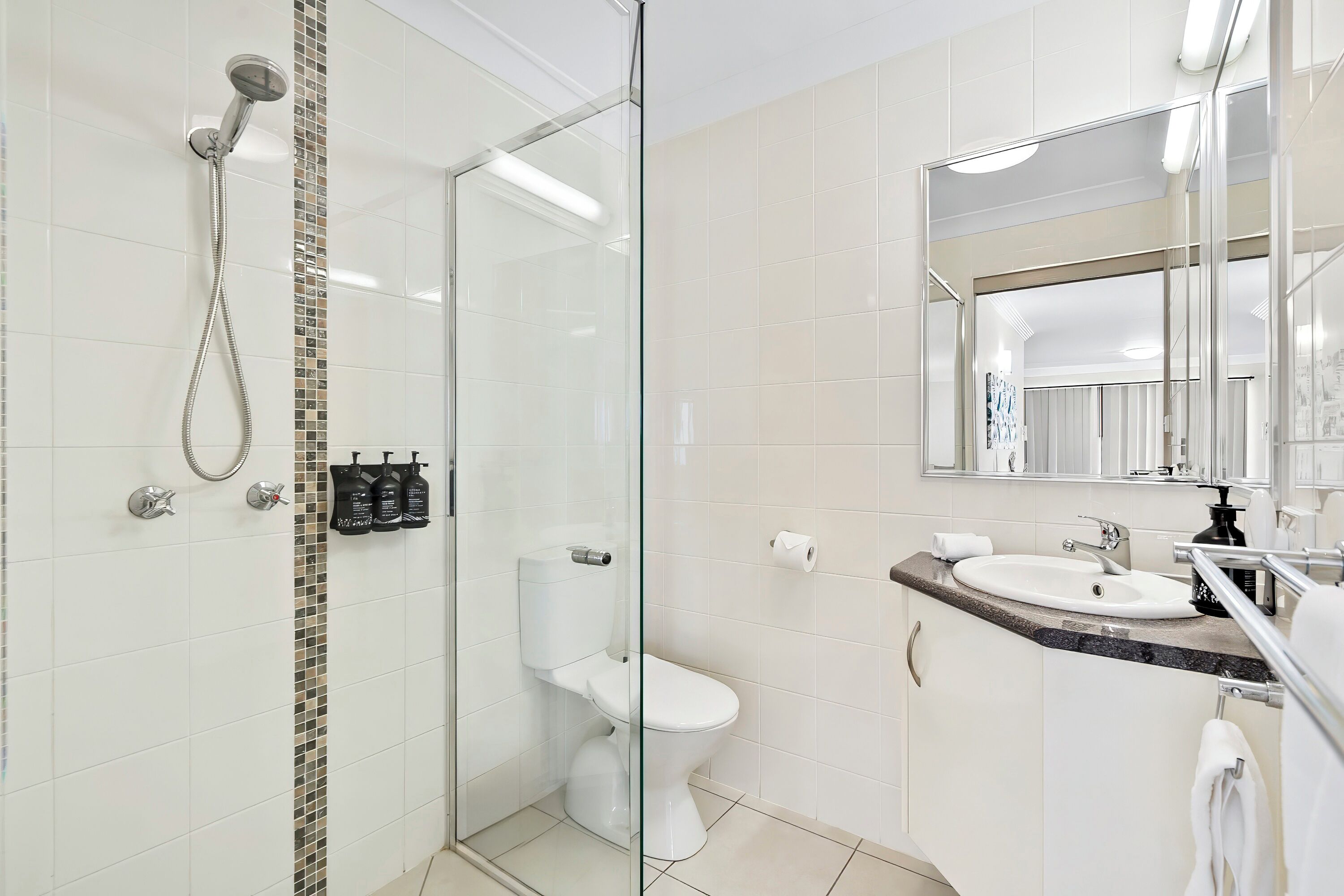 A modern bathroom with a glass shower enclosure, a white toilet, a sink with a mirror above, and a tiled wall.