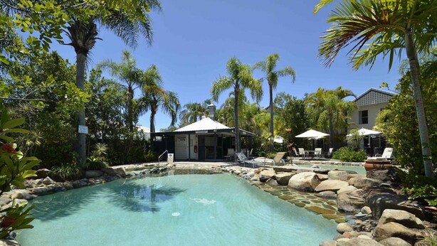 A pool area with lounge chairs and umbrellas, surrounded by rocks and palm trees. - Skippers Cove Waterfront Resort (Noosaville)