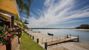 A beachside hotel with a wooden deck, a boat on the water, and a clear sky. - Skippers Cove Waterfront Resort (Noosaville)