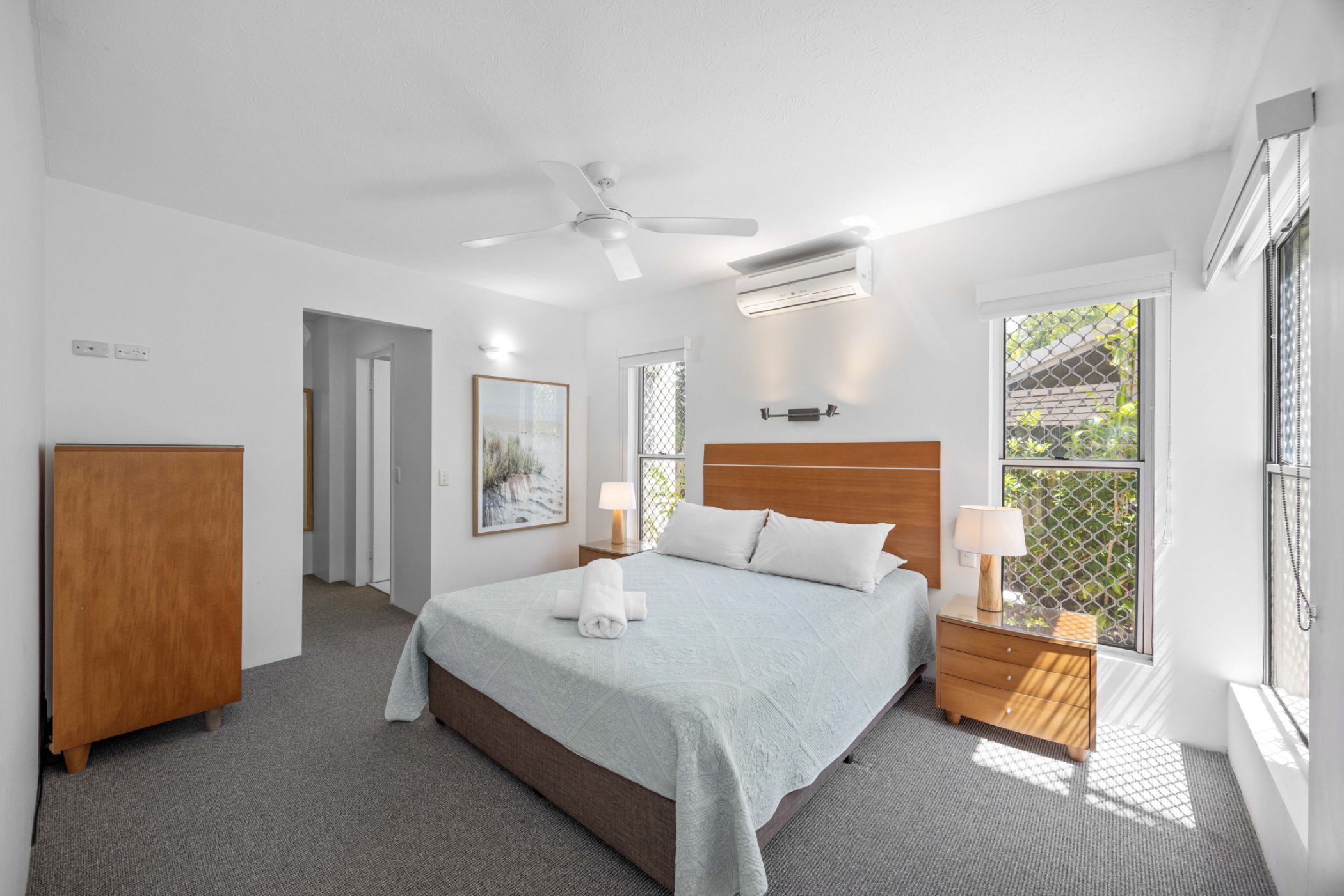 Een slaapkamer met een bed, nachtkastjes, een plafondventilator, een airconditioning en een raam met uitzicht, en een deur naar een andere kamer.