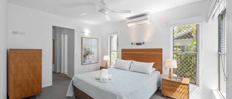 Een slaapkamer met een bed, nachtkastjes, een plafondventilator, een airconditioning en een raam met uitzicht, en een deur naar een andere kamer.