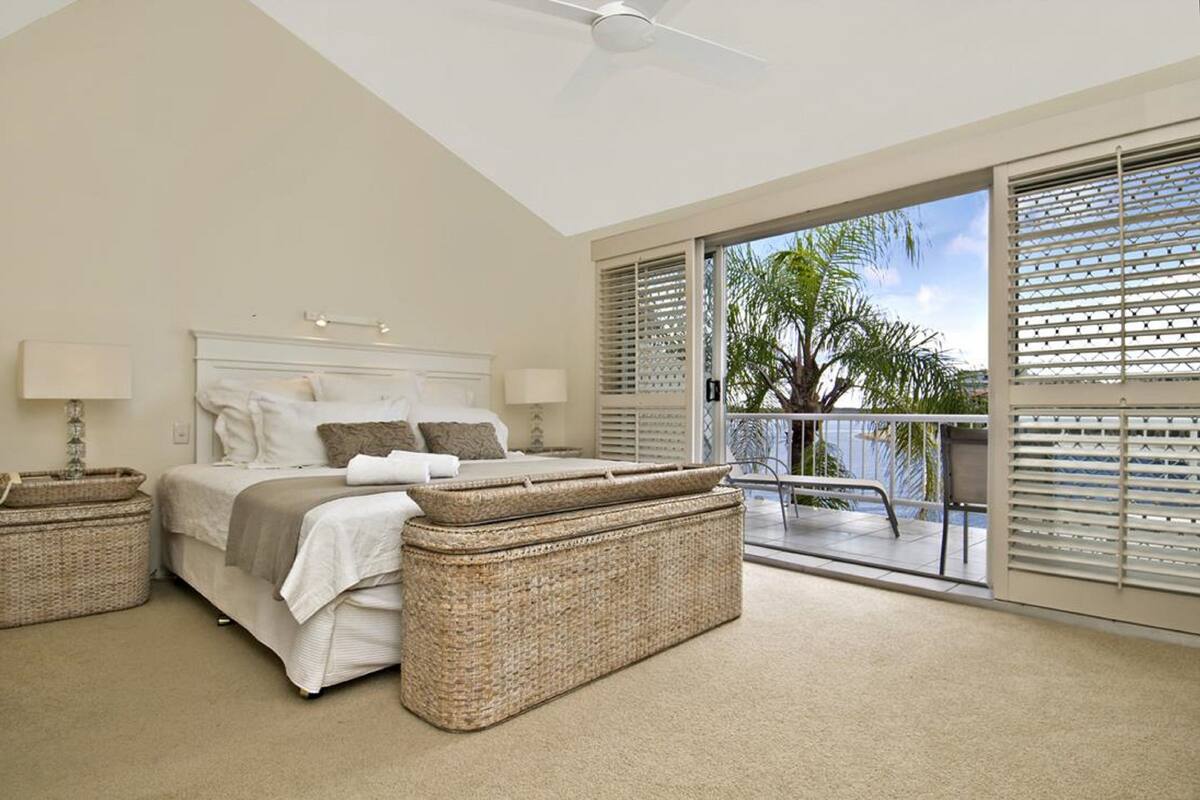 A bedroom with a bed, two nightstands, a ceiling fan, and a balcony with a view of palm trees.