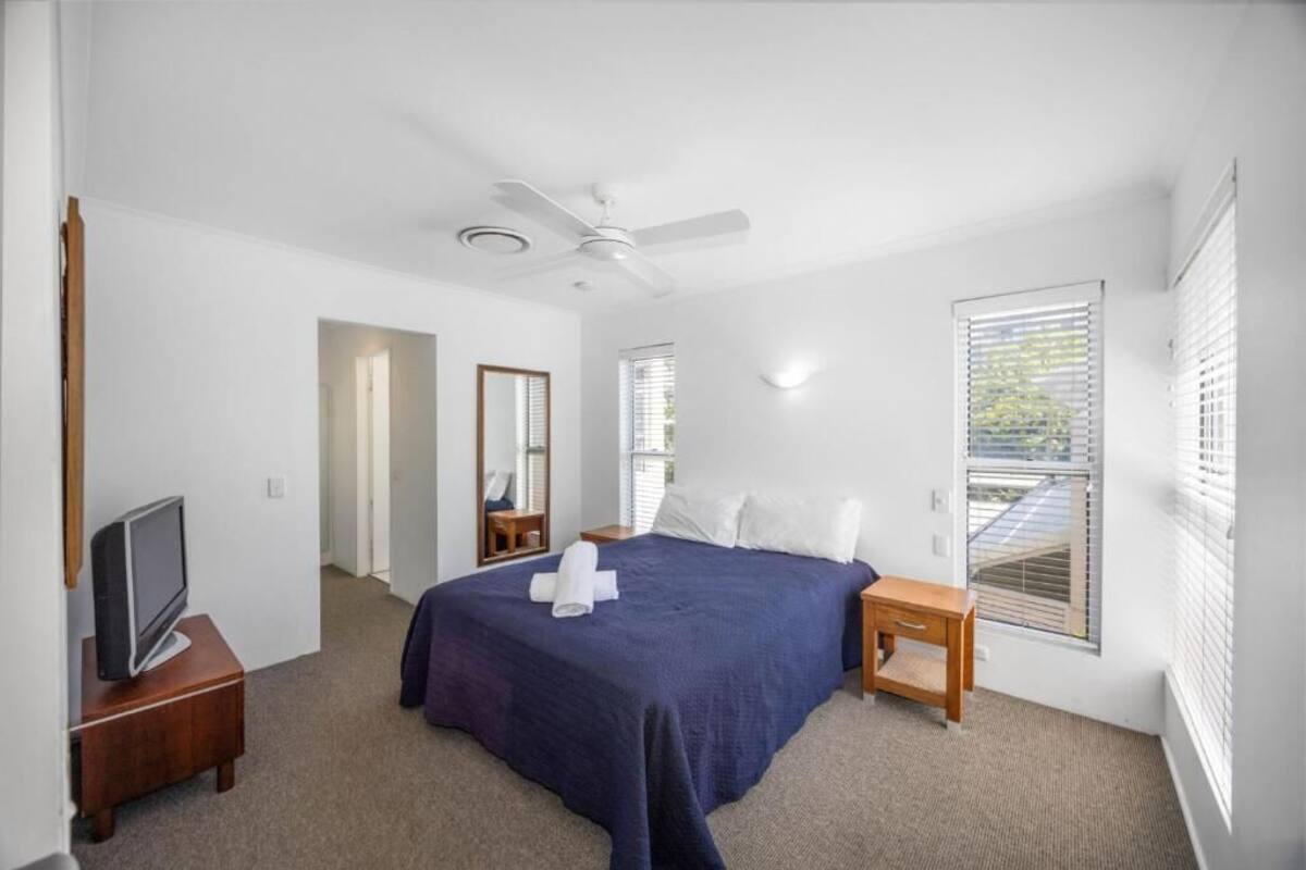 A bedroom with a bed, a television, a ceiling fan, and a window with blinds.