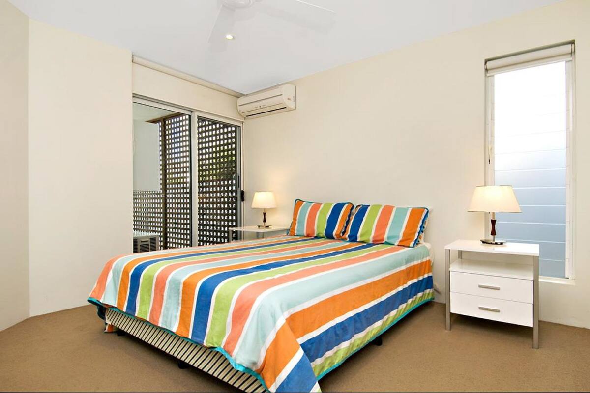 A bedroom with a striped bedspread, a white bedside table with a lamp, a window with blinds, and an air conditioning unit.