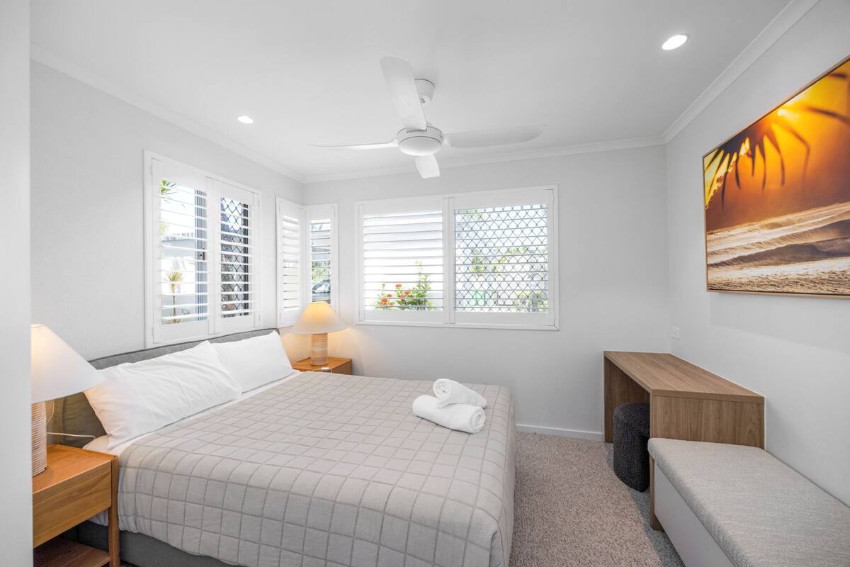 A bedroom with a bed, two bedside tables, a ceiling fan, a window with blinds, and a wall-mounted artwork.