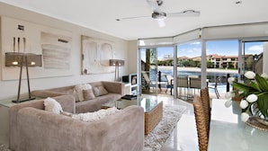 A modern living room with a large window, a ceiling fan, and a view of a pool and buildings. - Skippers Cove Waterfront Resort (Noosaville)