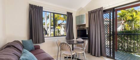 Una sala de estar con sofá, una mesa de comedor con sillas, televisor y un balcón con vista a palmeras.