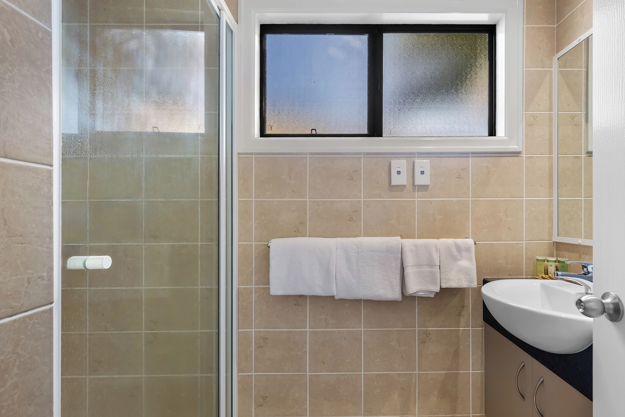 A bathroom with a glass shower enclosure, a window above the sink, and two towels hanging on a rack.