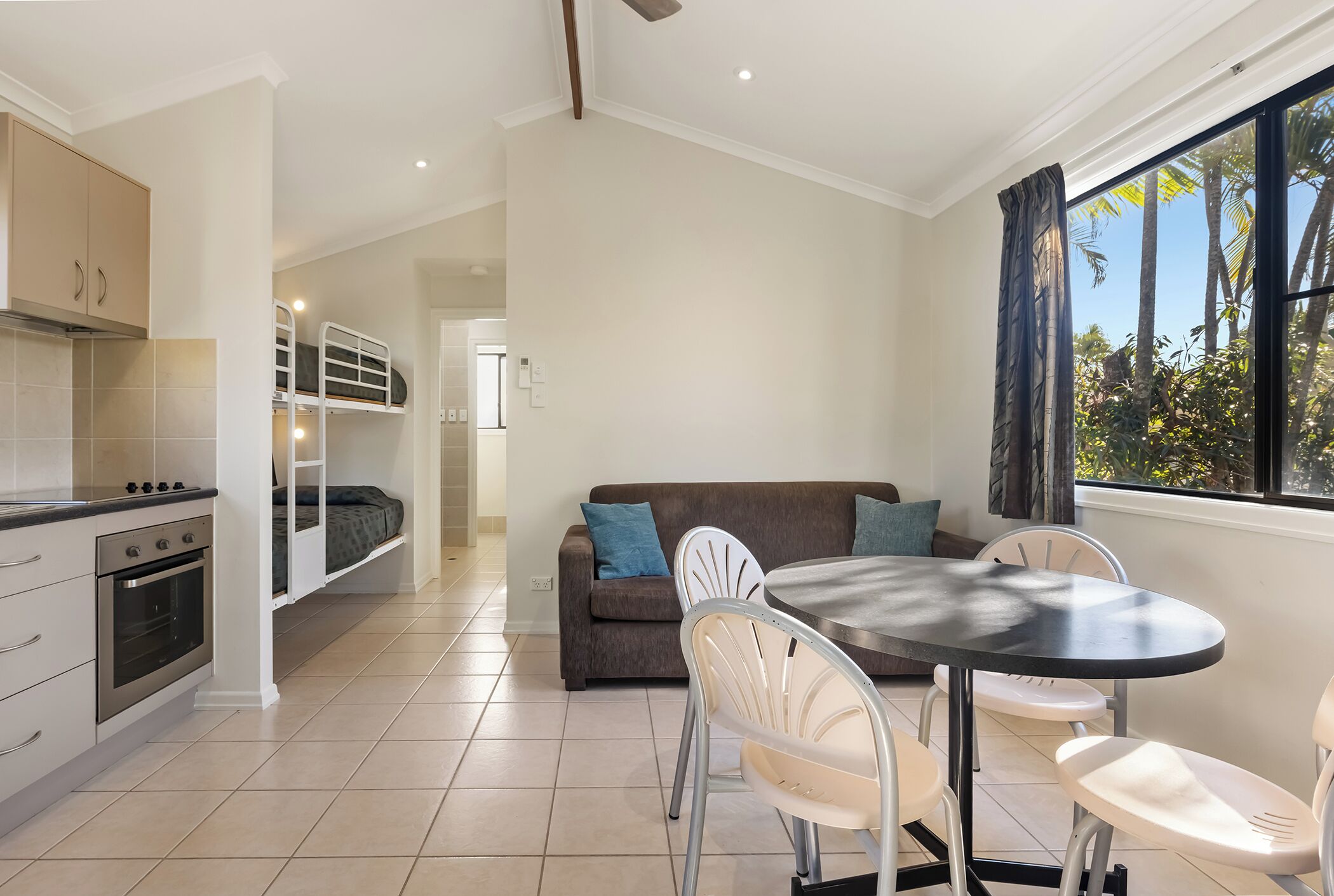 A compact kitchen with a built-in oven, a dining area with a round table and chairs, a sofa with cushions, and a large window with a view of palm trees.