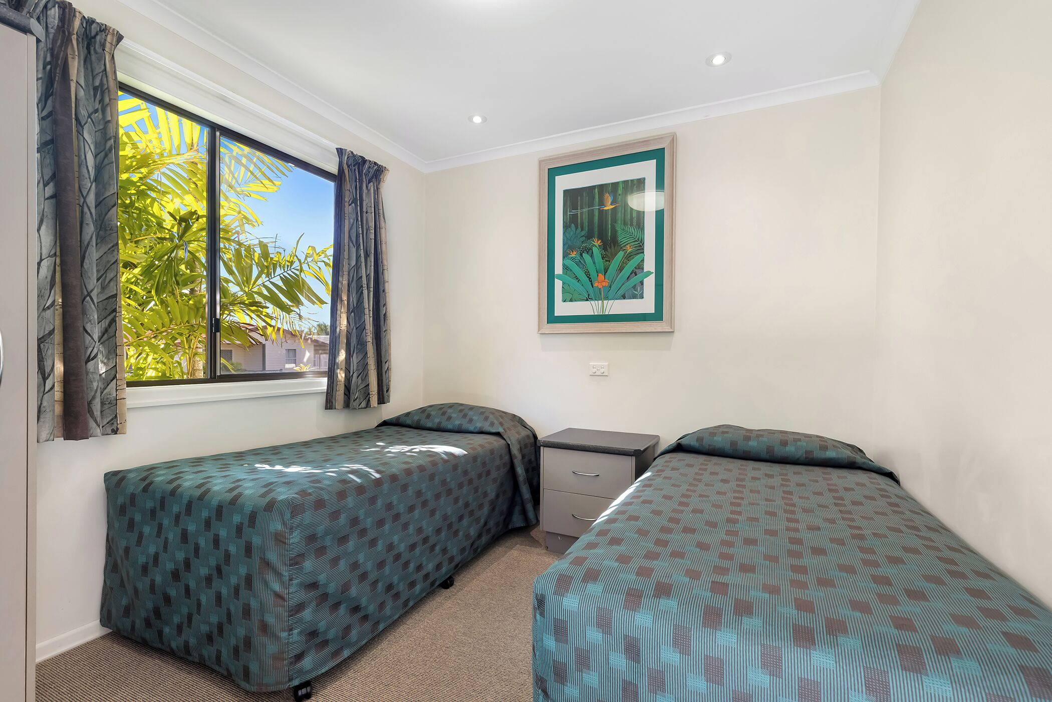 A room with two beds, a window with curtains, a small desk, and a framed picture on the wall.