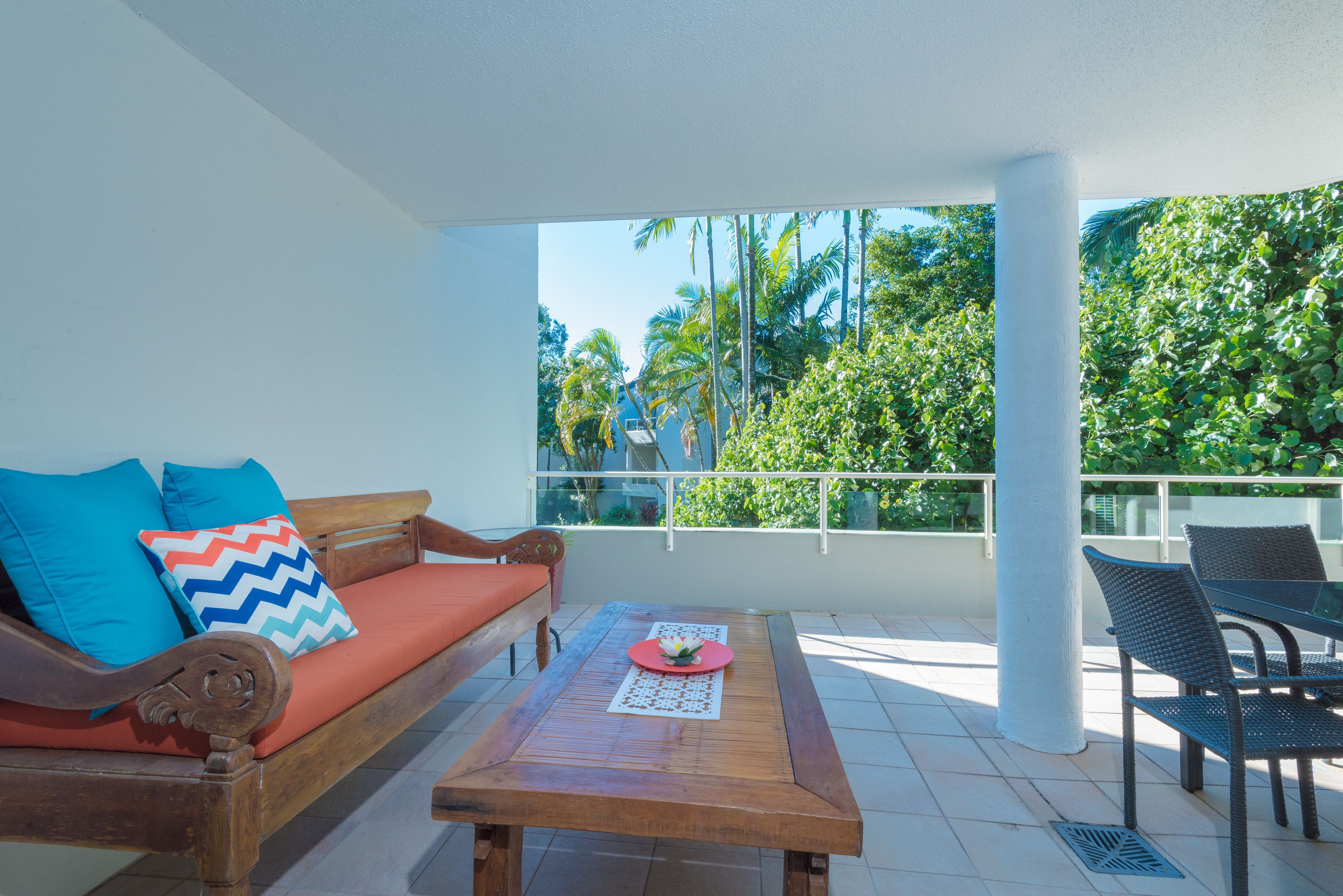 Ein Balkon mit Holztisch, einer Couch mit bunten Kissen und Blick auf üppiges Grün.