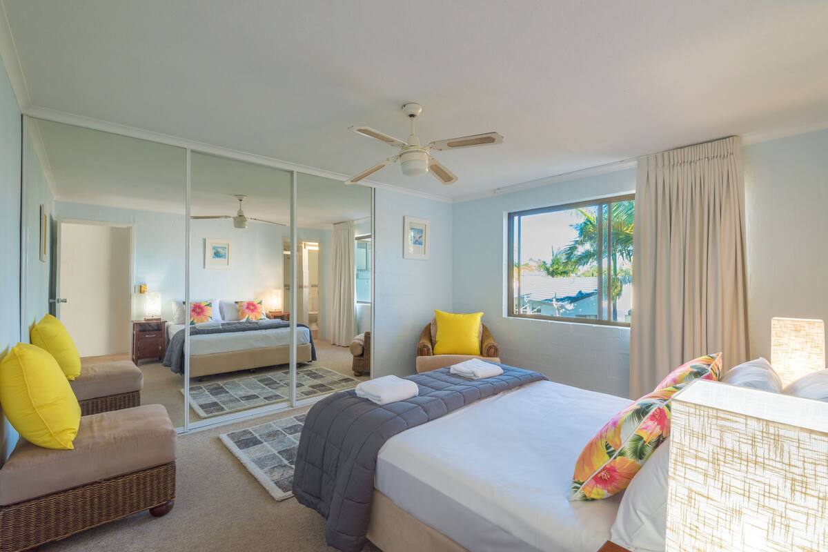 A hotel room with a bed, a chair, a mirror, a ceiling fan, and a window with curtains.