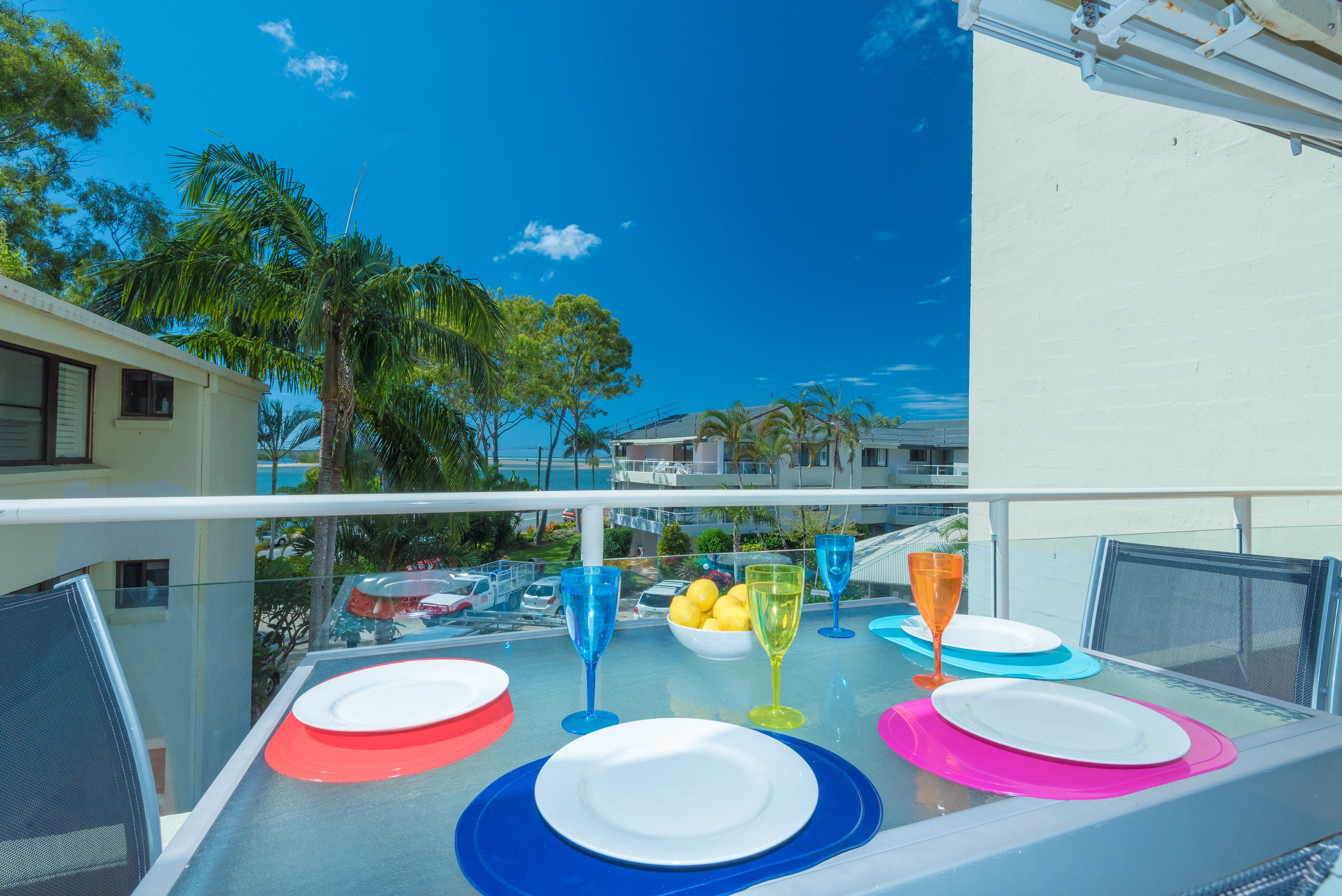 A balcony table set with colorful plates and glasses, overlooking a pool and palm trees.