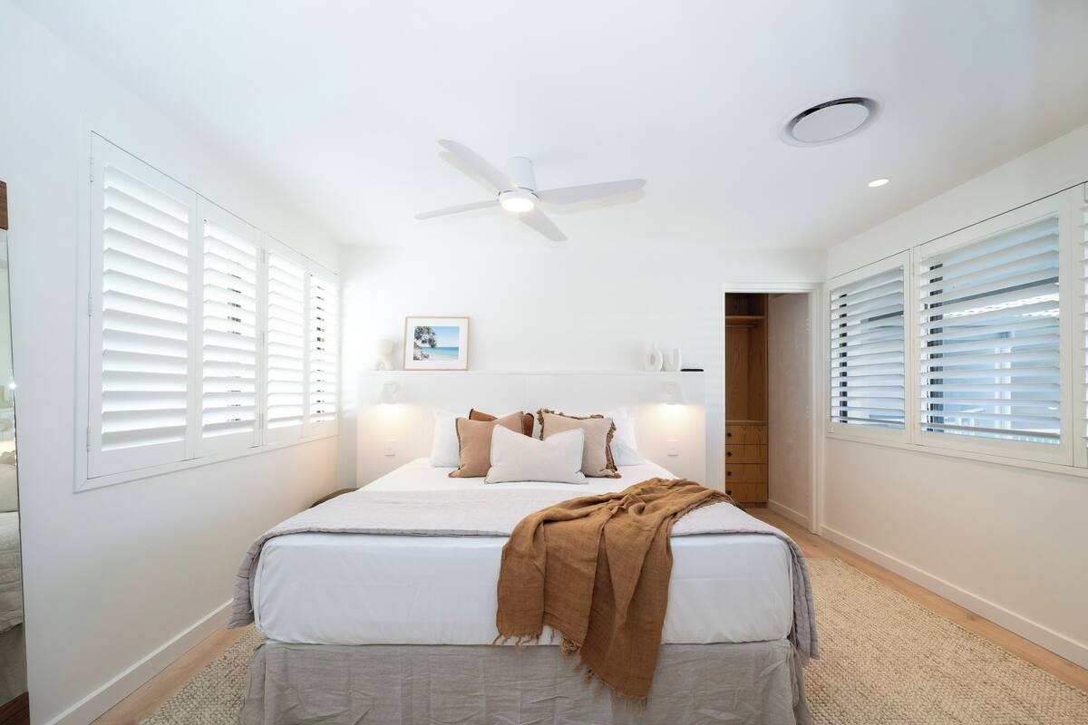 A bedroom with a bed, white walls, a ceiling fan, and a window with blinds.