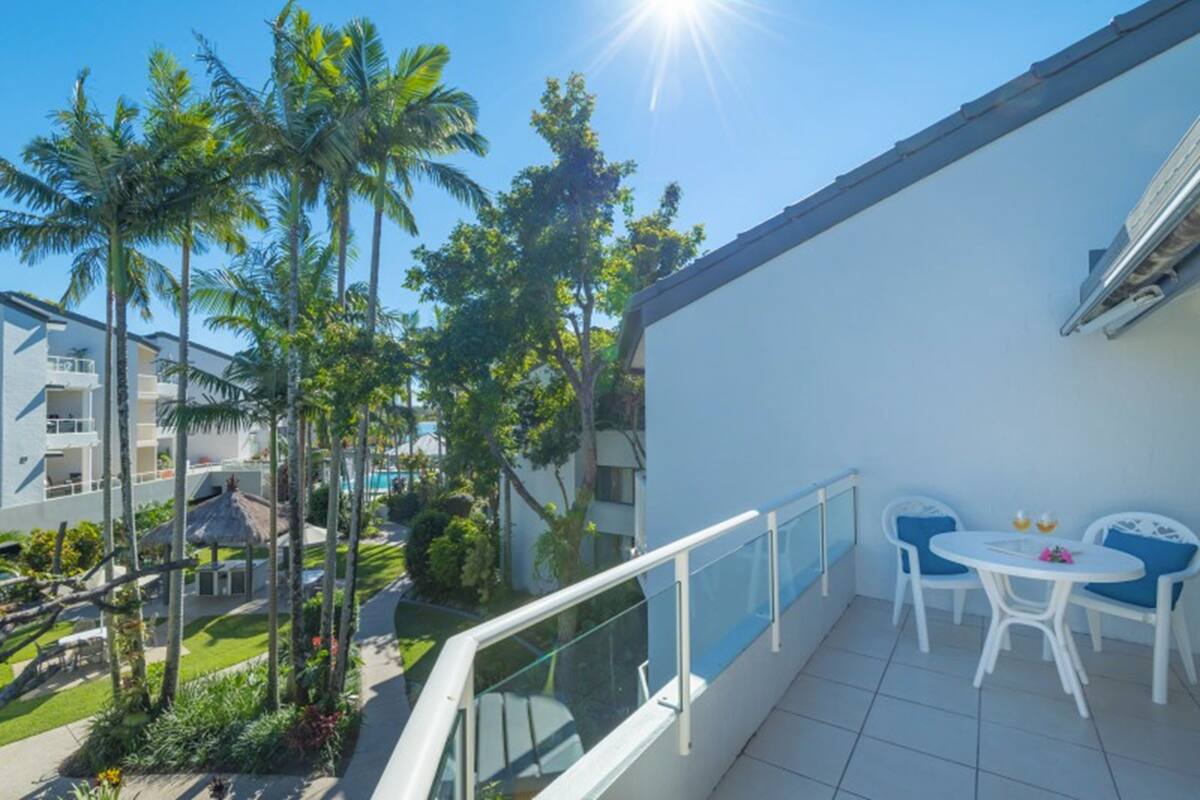A balcony with white chairs and a table, overlooking a pool and palm trees.