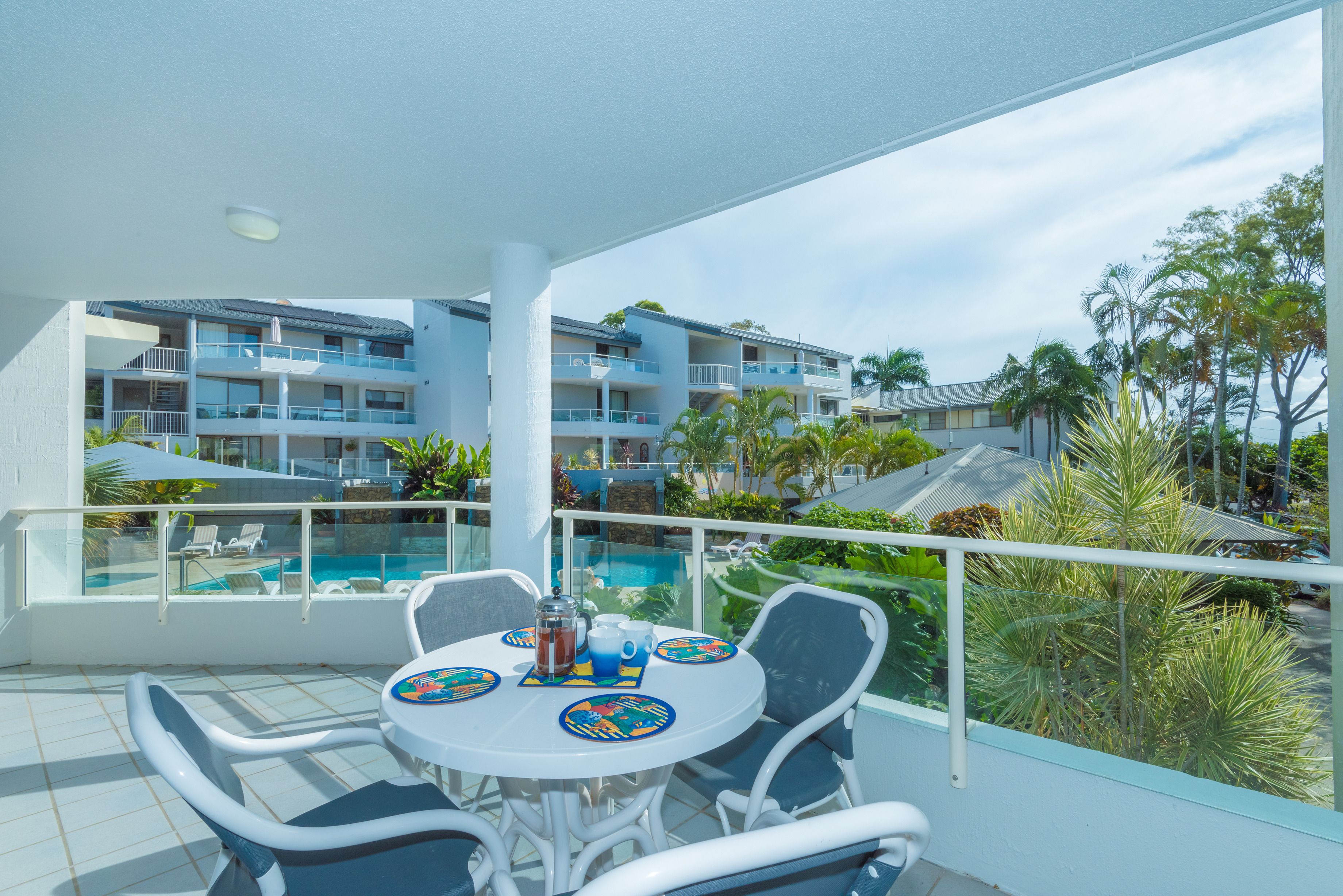 A balcony with a table, chairs, and a view of a pool and buildings.