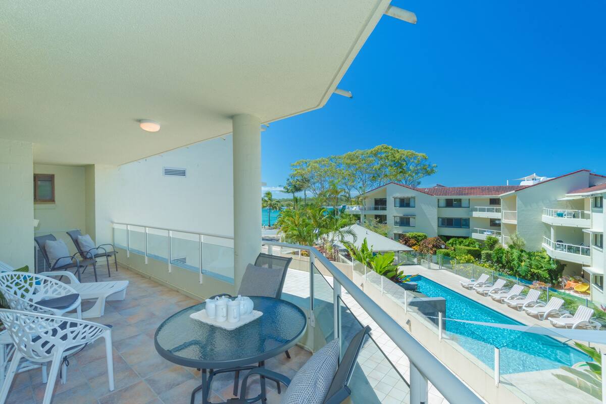 A balcony with a glass table, chairs, and a view of a swimming pool and buildings.
