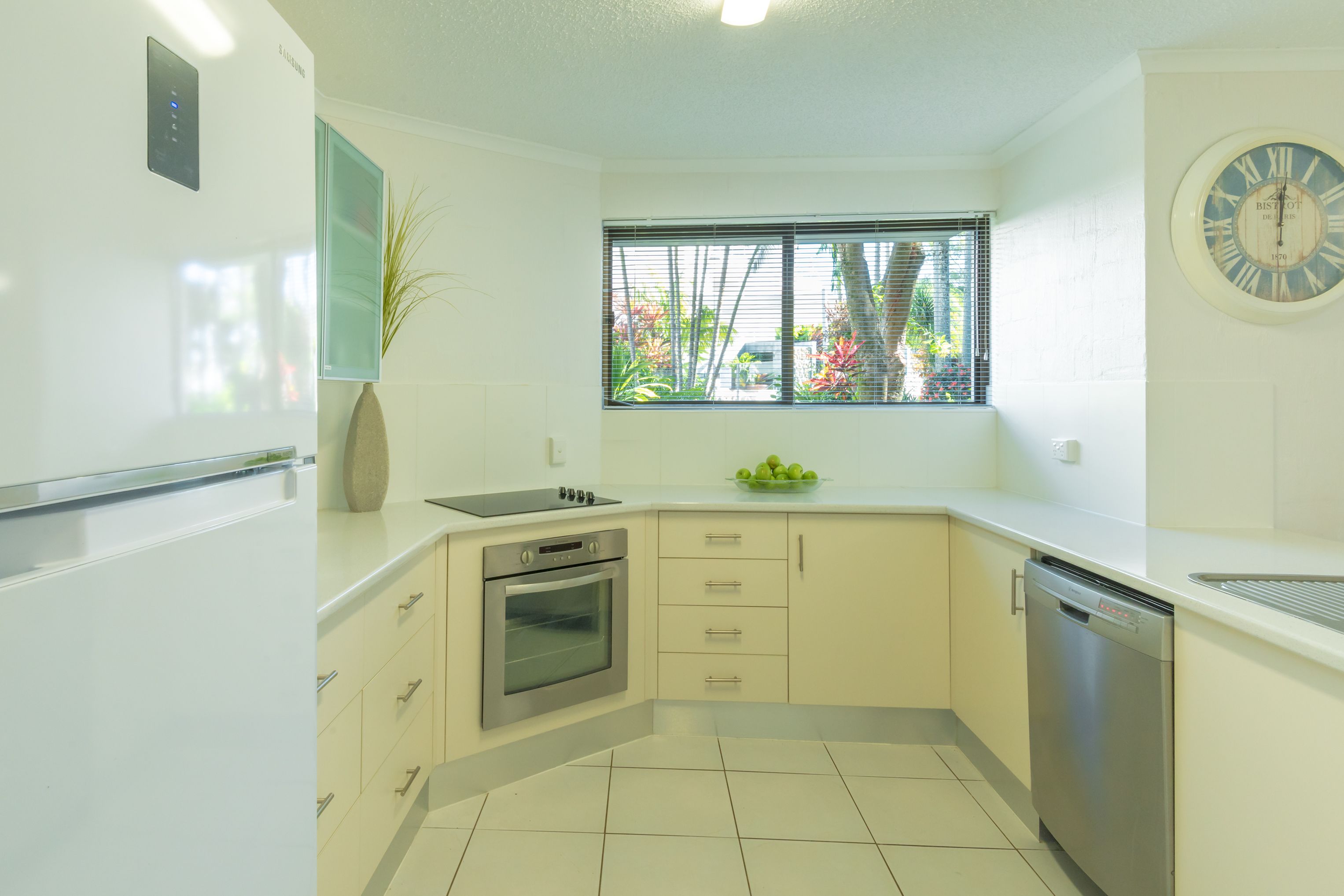 A modern kitchen with white cabinets, a built-in oven, a dishwasher, and a window with blinds.