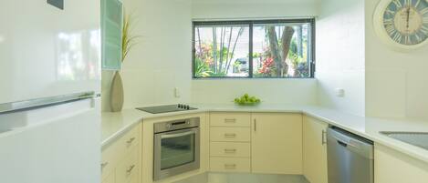 A modern kitchen with white cabinets, a built-in oven, a dishwasher, and a window with blinds.