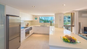 A modern kitchen with white cabinetry, stainless steel appliances, a large island with a fruit bowl, and a view of the outdoors through a window. - Munna Beach Apartments (Noosaville)