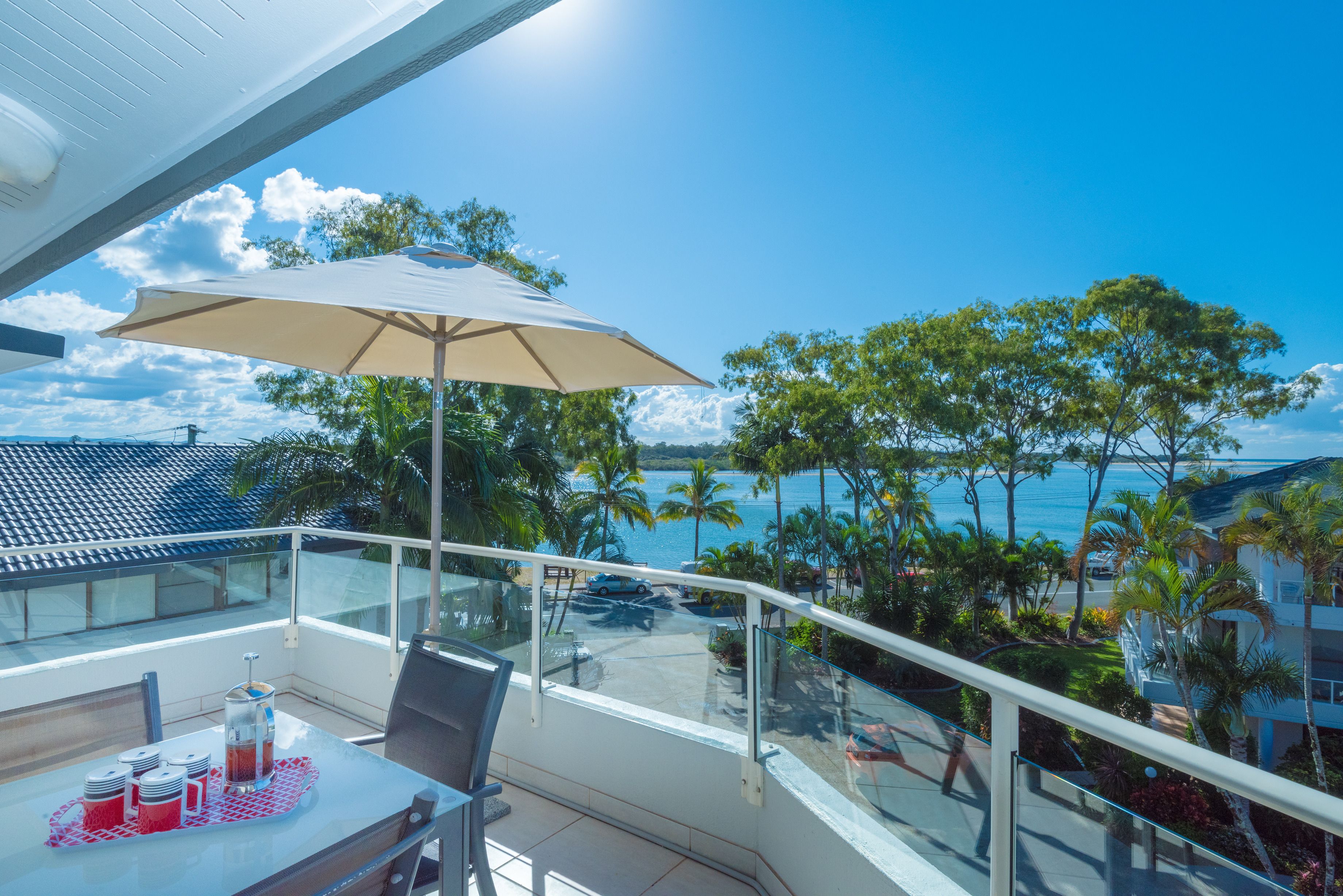 Varanda com mesa, cadeiras e guarda-sol, com vista para uma área costeira com palmeiras e o mar.
