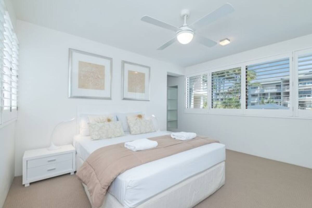A bedroom with a bed, nightstand, ceiling fan, and large windows.