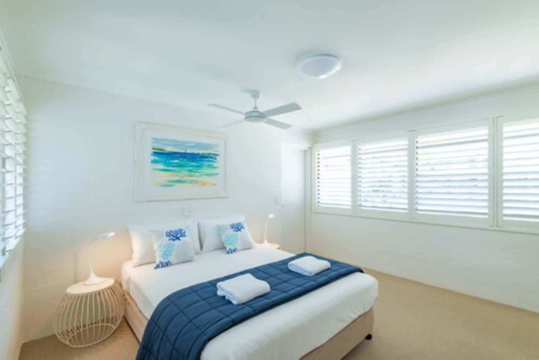 Un dormitorio con cama, ventilador de techo, ventana con persianas y una obra de arte colgada en la pared.