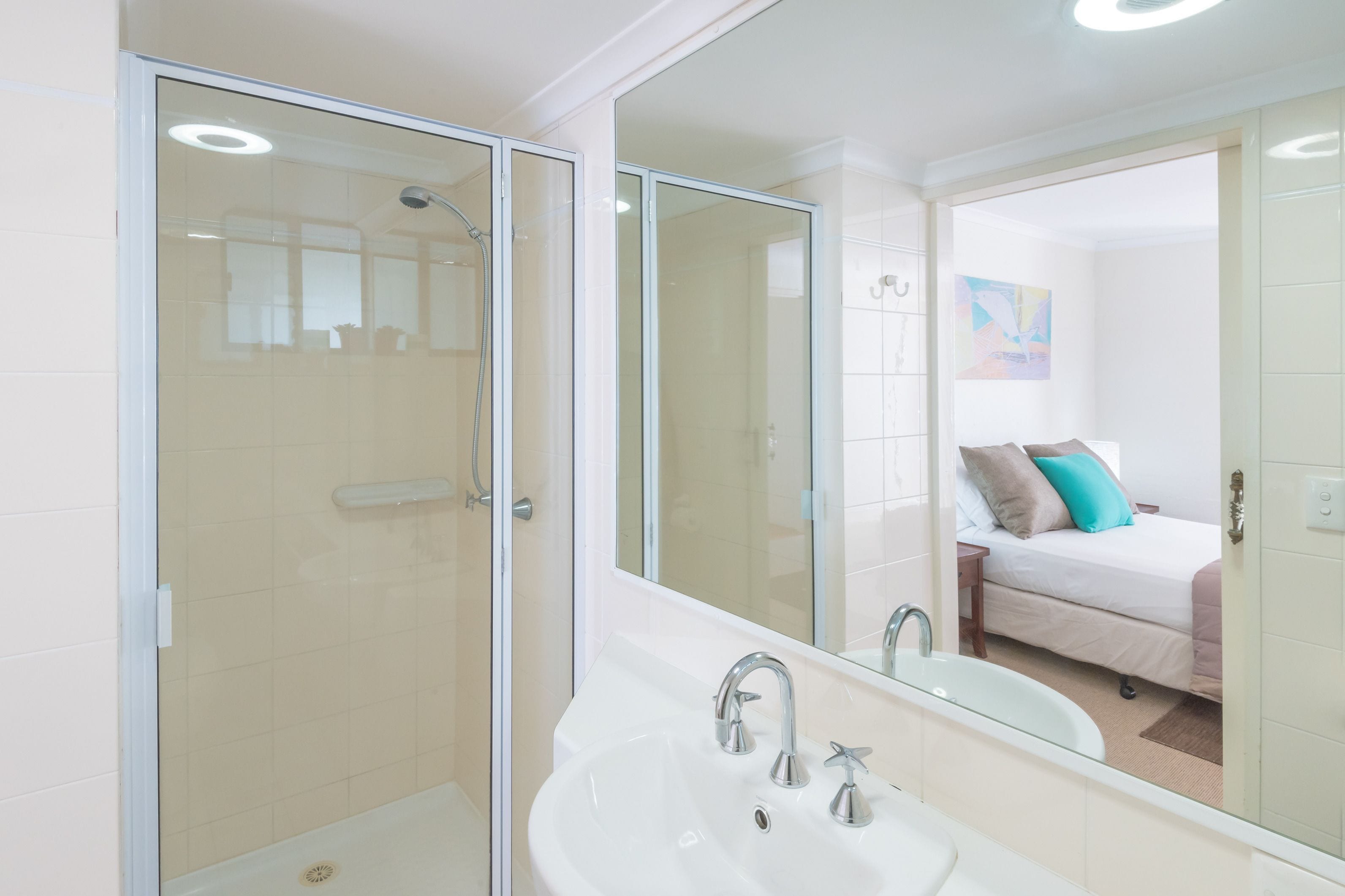 A modern bathroom with a glass shower enclosure, a white sink, and a large mirror reflecting a bedroom with a bed and pillows.