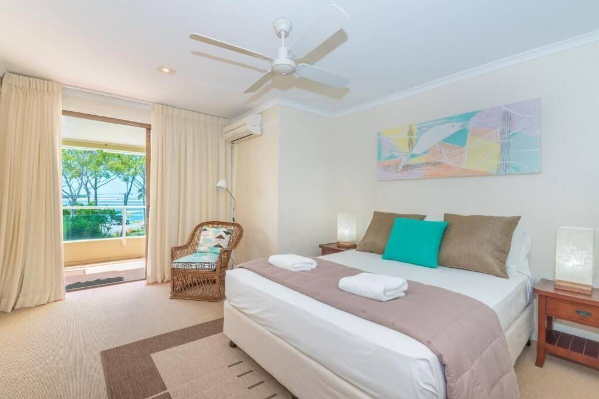 A hotel room with a bed, a ceiling fan, a chair, a nightstand, and a painting on the wall.