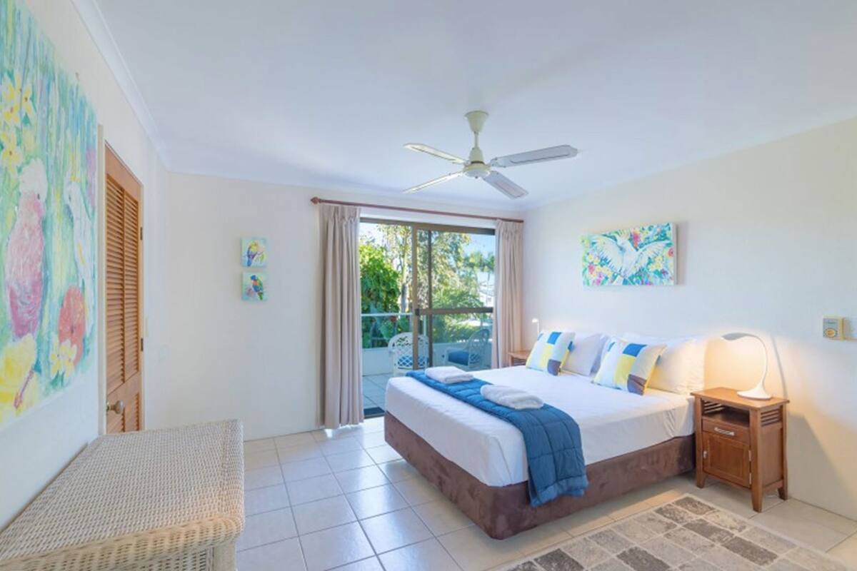 A bedroom with a bed, a ceiling fan, a nightstand, a lamp, and a view of the outdoors.