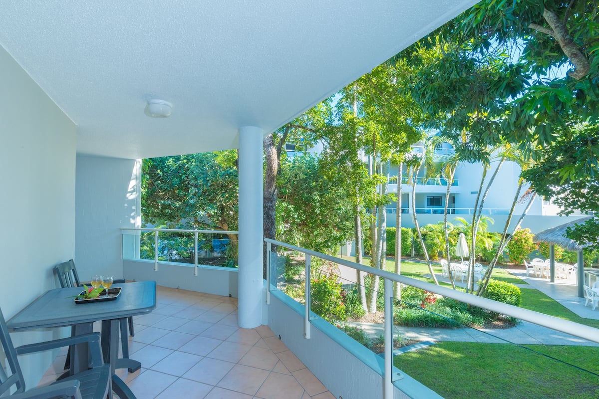 A balcony with a table and chairs, a railing, and a view of a garden and buildings.
