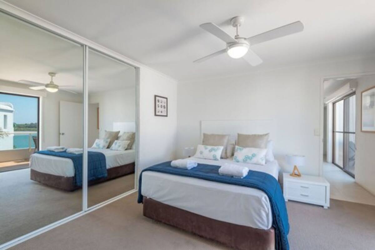 A bedroom with two beds, a ceiling fan, a large mirror, and a view of the outside.