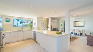 A modern kitchen with white cabinetry, a central island, and a dining area with a table and chairs. - Munna Beach Apartments (Noosaville)