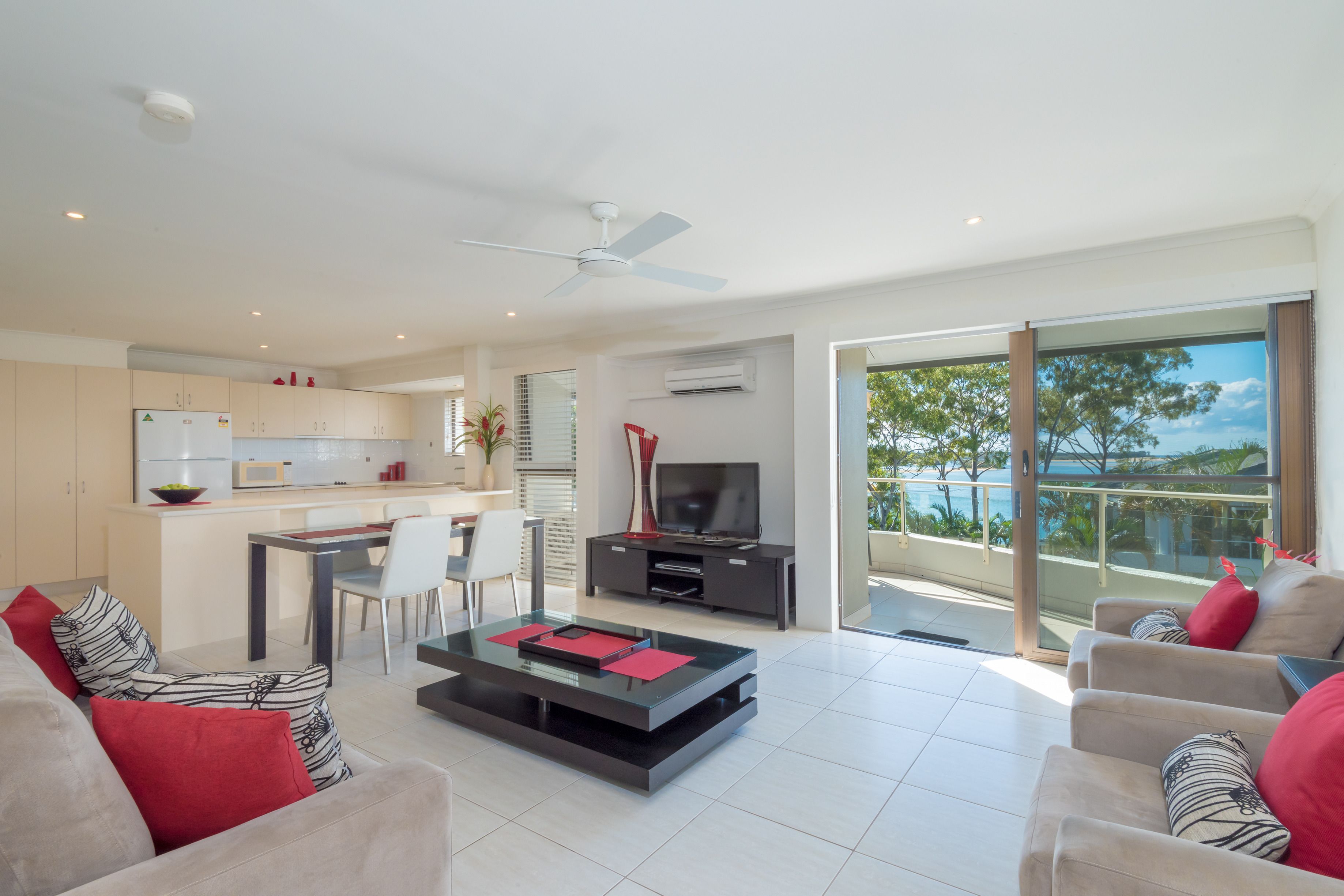 A modern living room with a sofa, coffee table, and a television. There is a kitchen area with a refrigerator and a dining table. The room has large windows and a balcony with a view of trees.